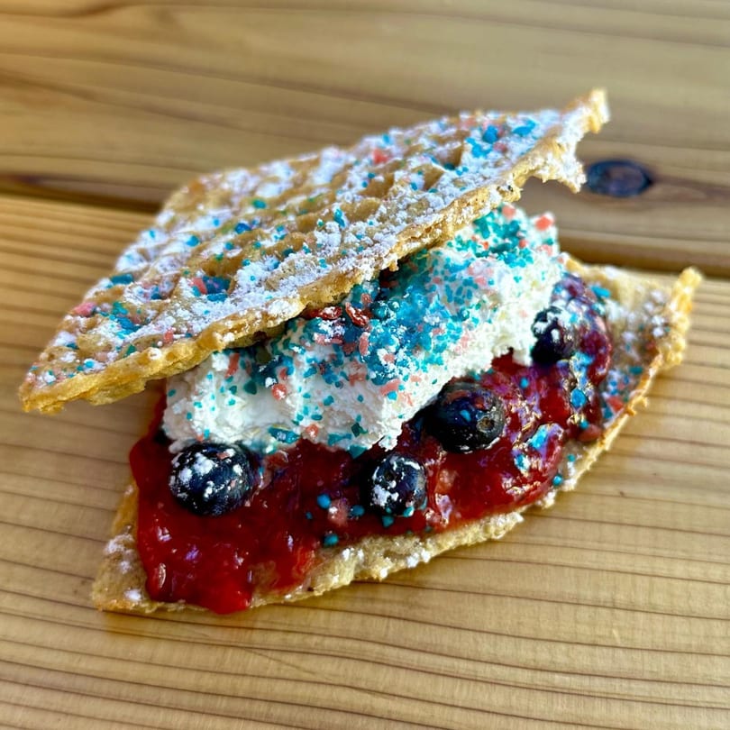A Smaaken waffle sandwich with red jam, blueberries, whipped cream, and colorful sprinkles.