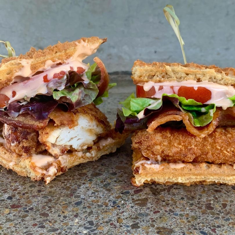 Two Smaaken Waffle Sandwiches with fried chicken, bacon, lettuce, tomato, and sauce on a speckled surface.