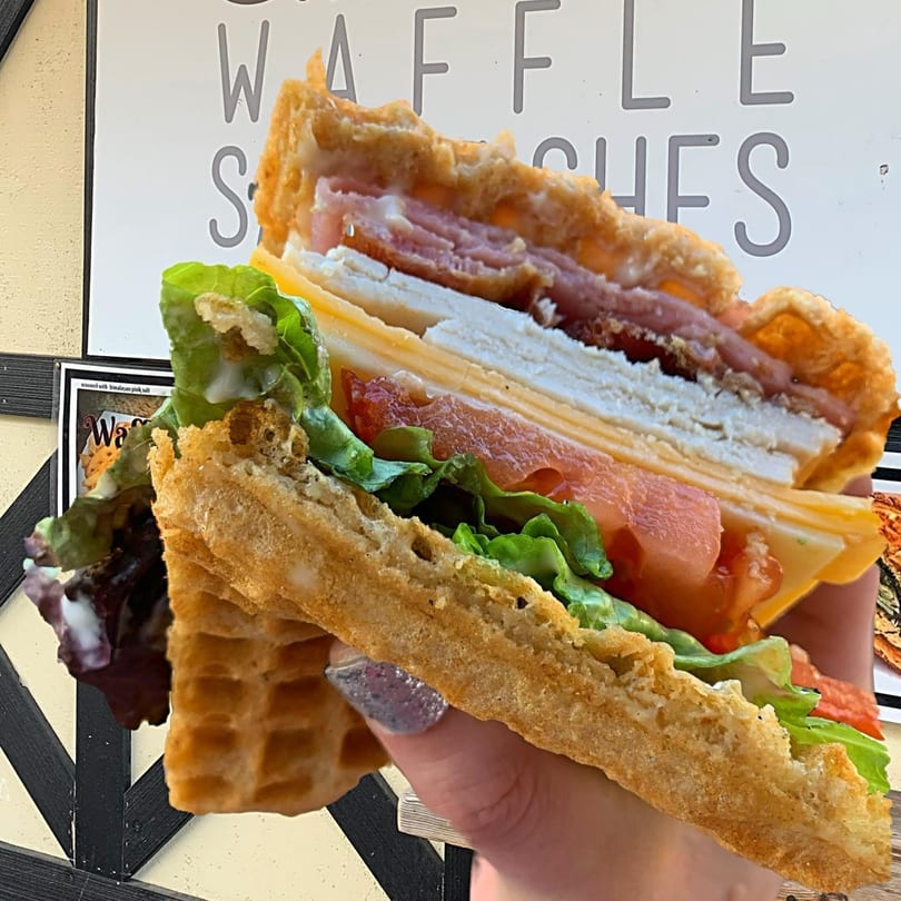 Hand holding a Smaaken Waffle Sandwich filled with ham, turkey, cheese, lettuce, and tomato.