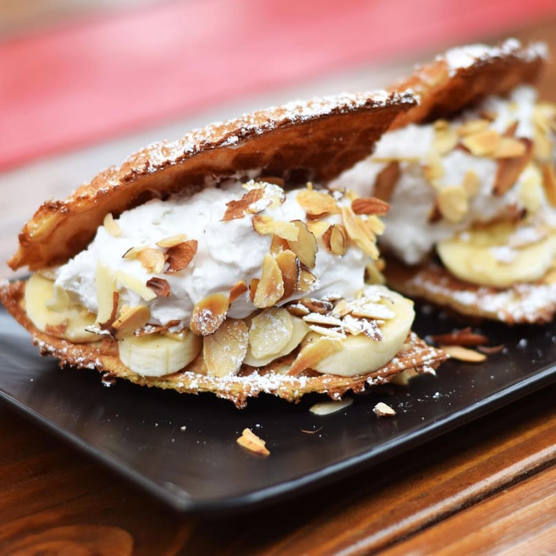 Two Smaaken Waffle Sandwiches filled with whipped cream, sliced bananas, and almonds, dusted with powdered sugar.