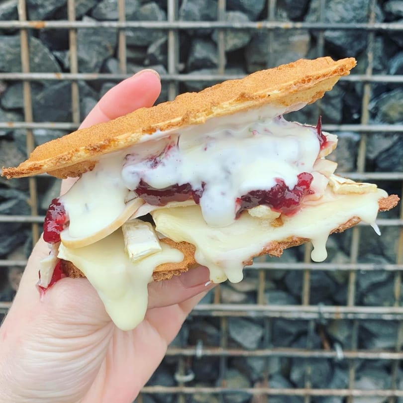 Hand holding a Smaaken waffle sandwich with melted brie, cranberry sauce, and white cream.
