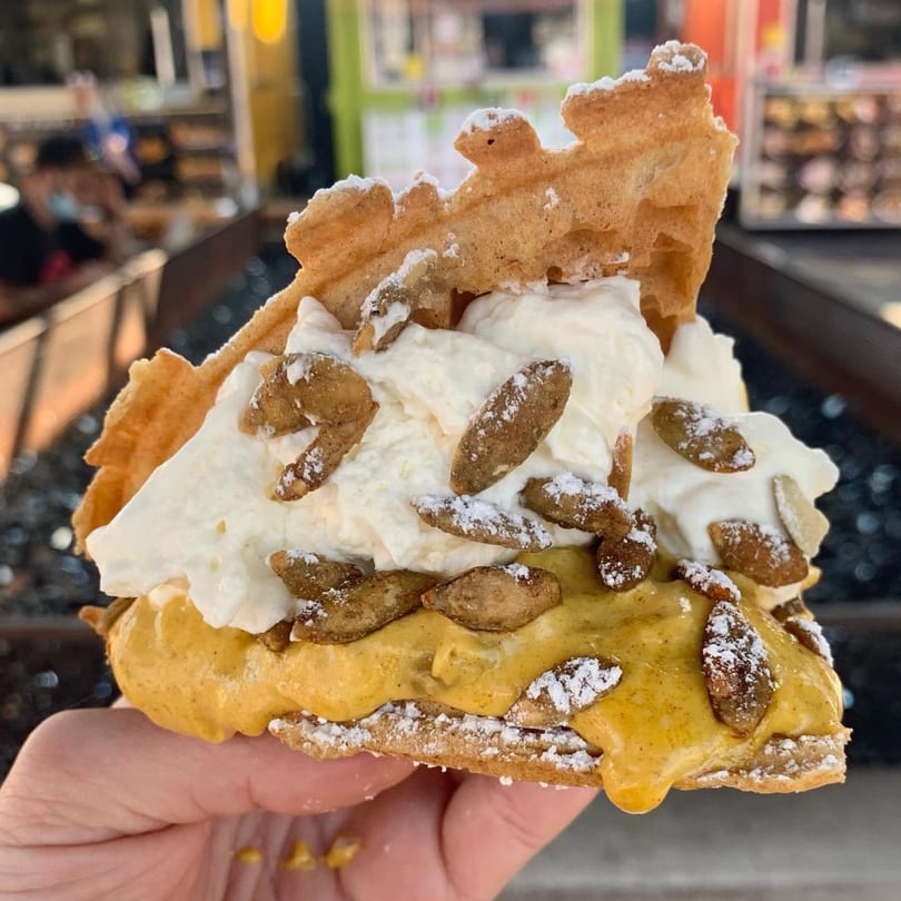 A hand holds a Smaaken waffle sandwich with pumpkin cream, whipped cream, and candied pumpkin seeds.