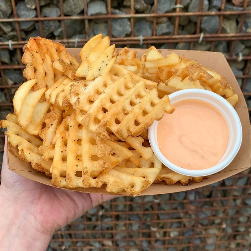 A hand holds a cardboard tray of Smaaken waffle fries with a side of dipping sauce.