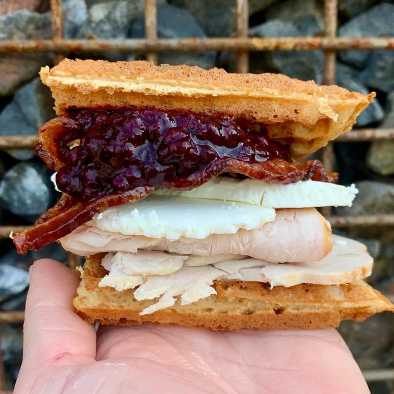 Hand holding a Smaaken waffle sandwich with turkey, cheese, bacon, and berry jam.